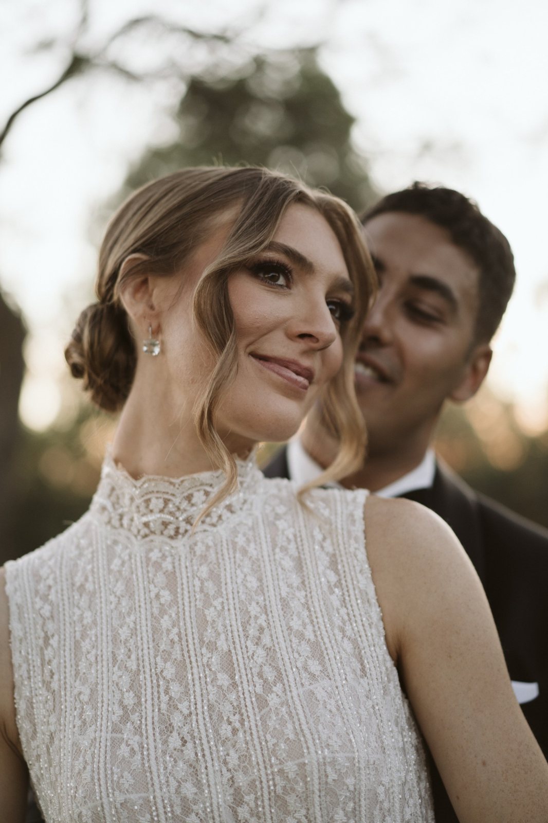 Bride with a low chignon and lace high-neck dress smiling at golden hour, groom behind her
