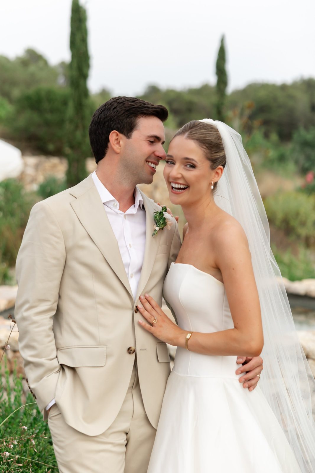 Laughing bride with a sleek updo and veil beside the groom in a beige suit, Mediterranean garden